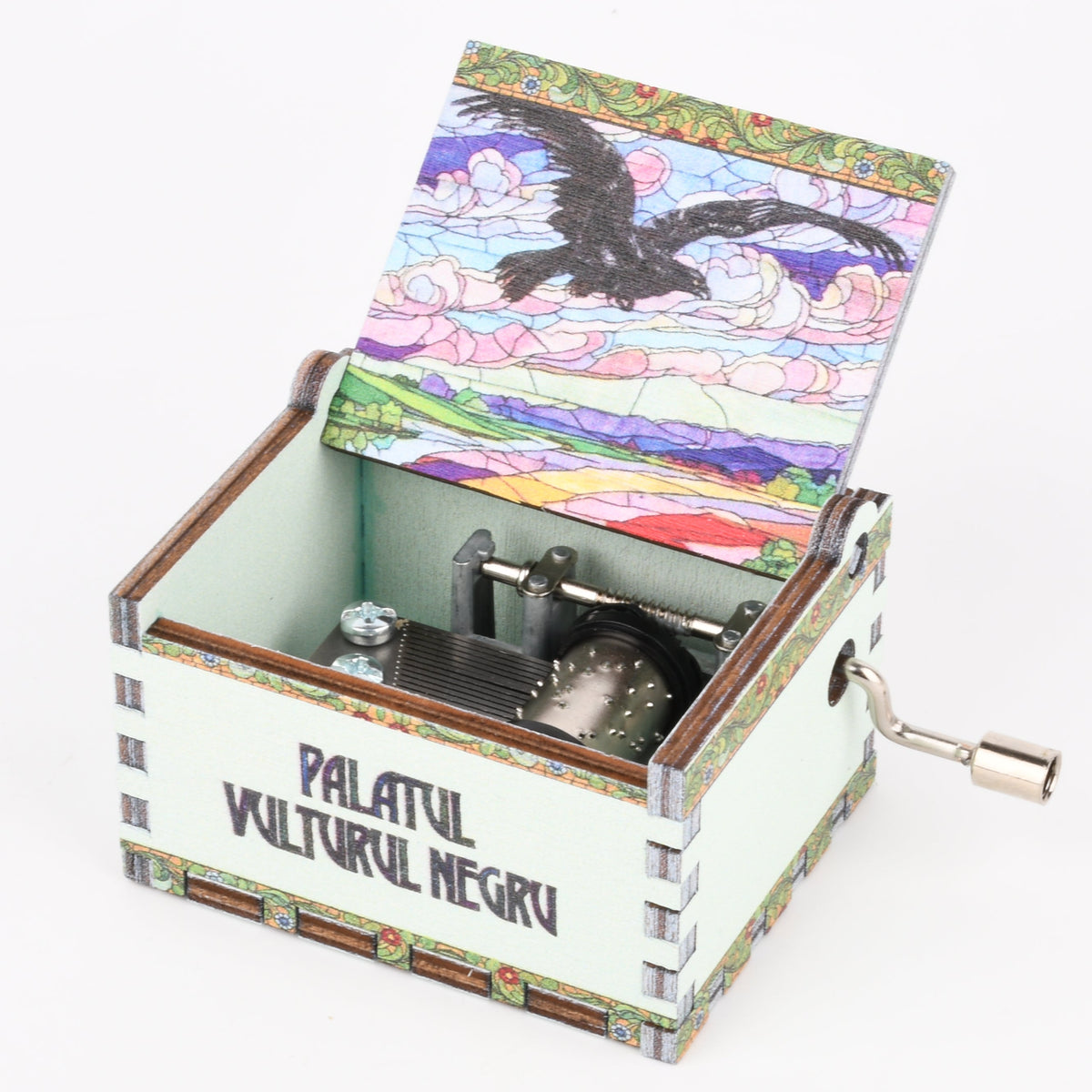 A hand-cranked music box with a white body and a colorful lid illustration of a black bird over a landscape, featuring the text 'PALATUL VULTURUL NEGRU' on the front, set against a plain white background.
