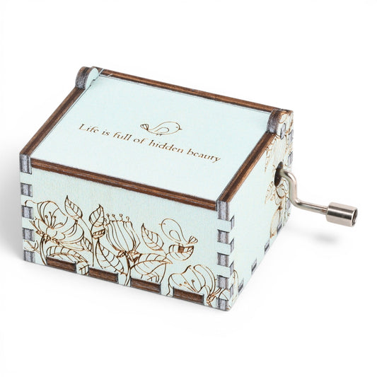 Enchanted Music Boxes hand-crank music box in light blue plywood with floral engravings and a metal crank handle.