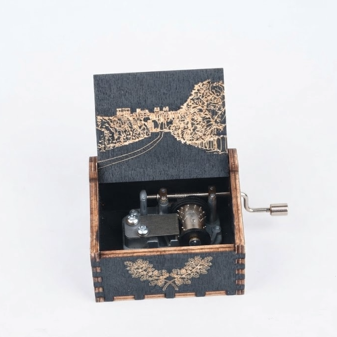 A wooden music box with a dark gray textured exterior and light brown laser-engraved details, including a landscape design on the lid and a floral motif on the front. The box is open, showing the internal mechanism, and has a silver metal crank handle on the right side, set against a plain white background.